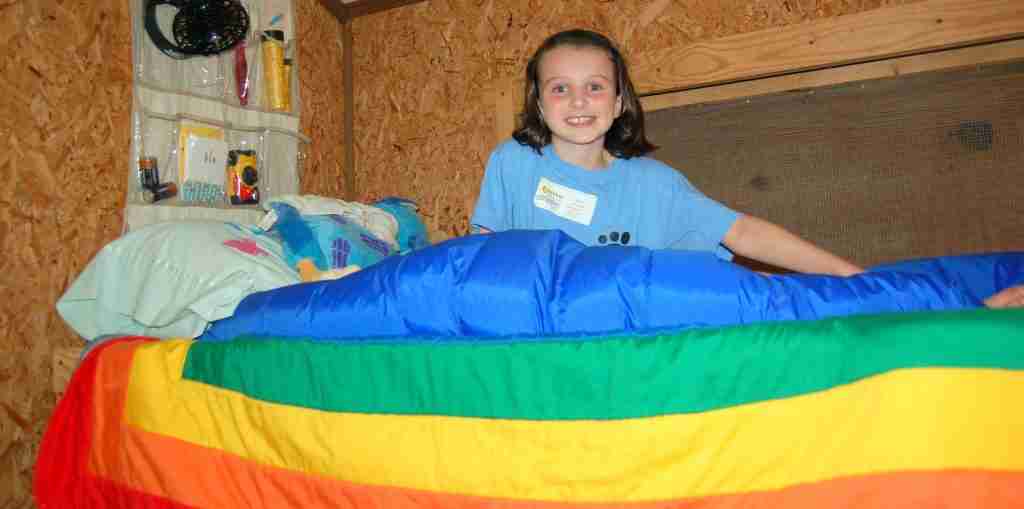 A young camper smiling from her bunk with a rainbow sleeping bag