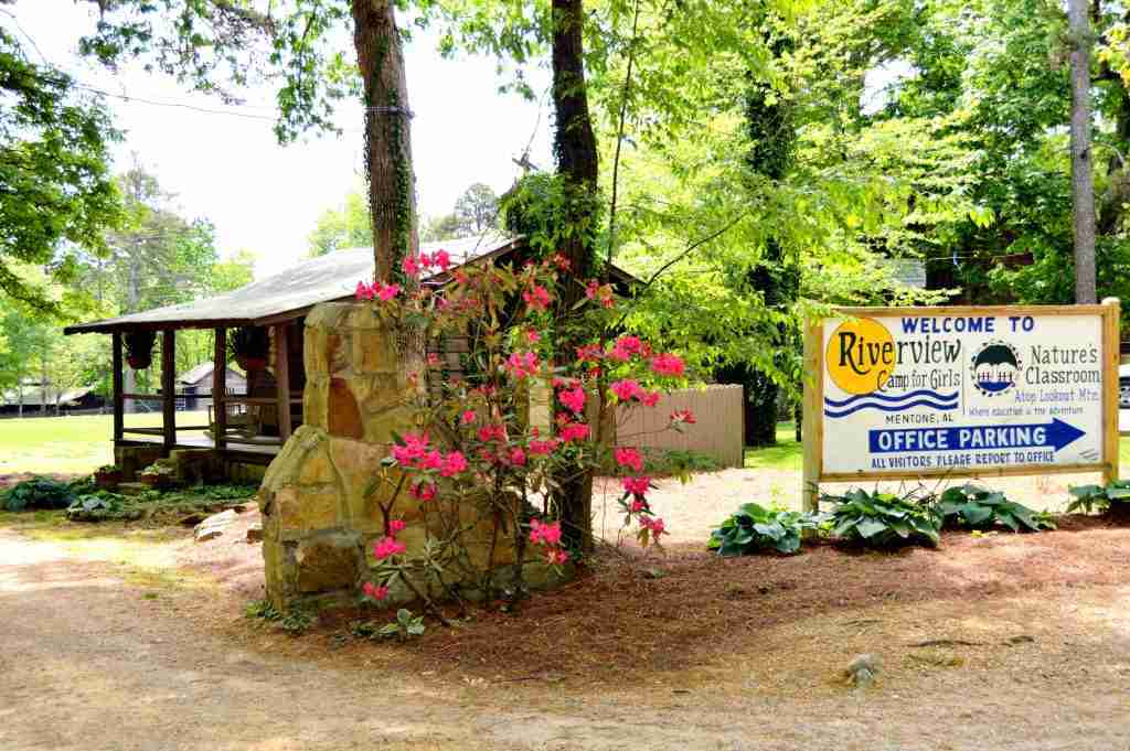 The Riverview Camp for Girls welcome sign beside a stone pillar and blooming rhododendron